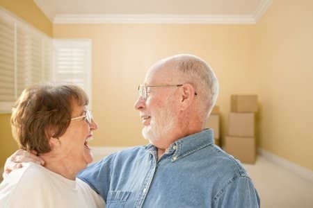 Elderly couple with glasses is looking at each other with joy with moving boxes in the far-right hand side
