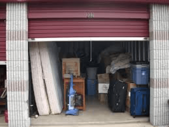 Maroon colored storage unit filled with personal belongings, two mattresses, blue vacuum, suitcases