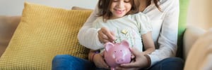 Lady with her daughter sitting on a couch holding a pink piggy bank